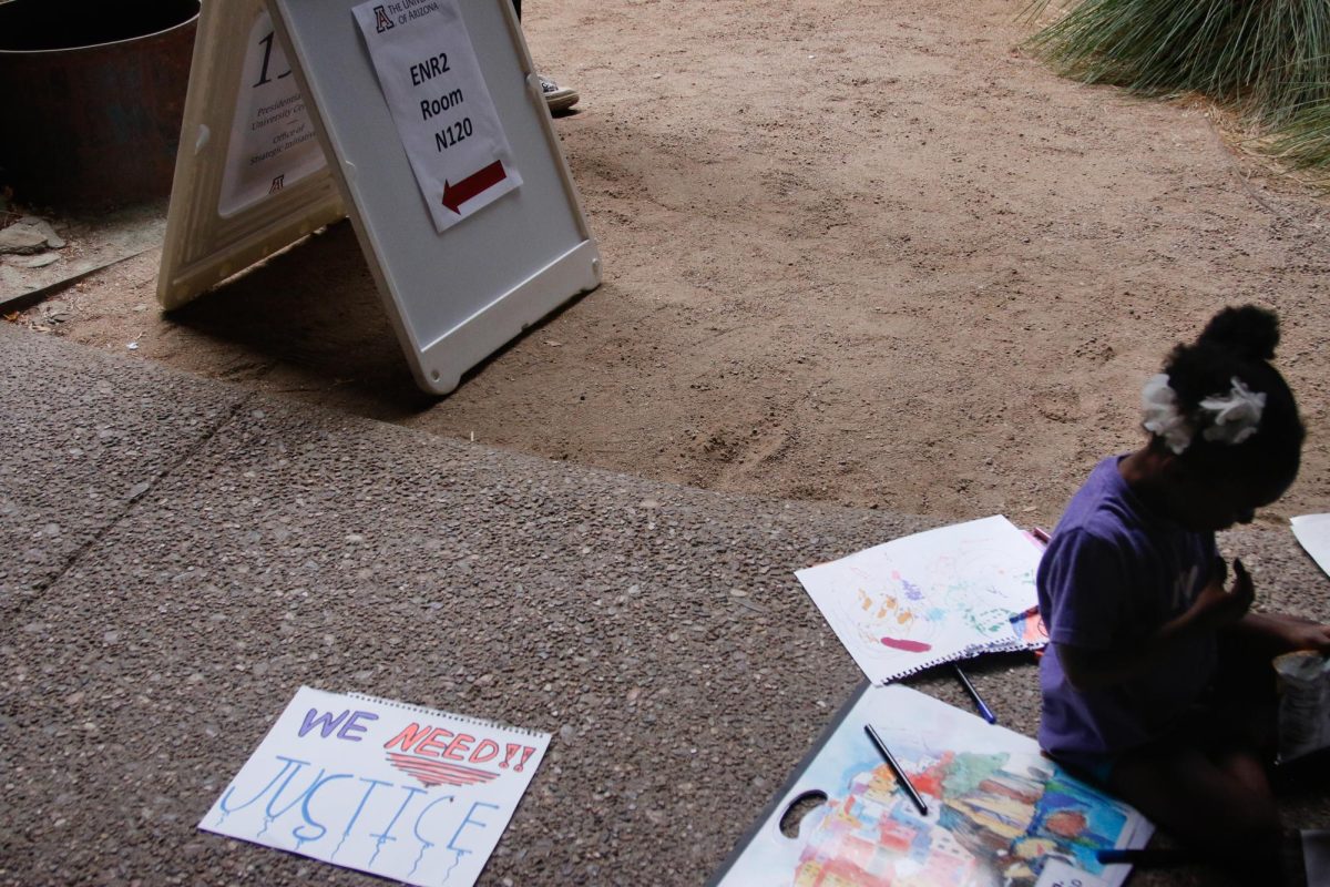 A child sits outside the Arizona Board of Regents town hall meeting on April 24, working on outlining letters on poster board next to a completed protest sign that reads “We Need Justice.” The town hall was held on the University of Arizona campus at the Environment and Natural Resources 2 building. 